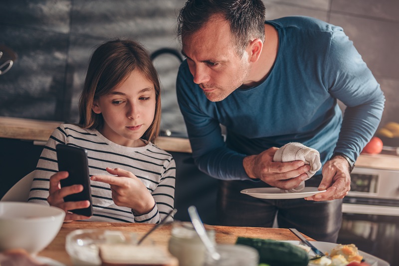 teenage child indicating to their mobile phone screen, father drying a dish in the kitchen in the background looks toward the phone