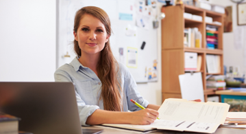 This image shows a female teacher sitting at her desk in a bright classroom.