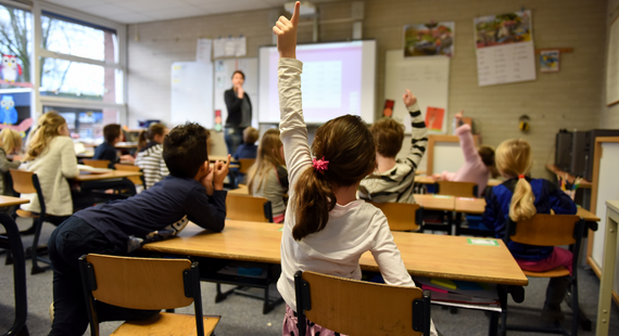 A young girl raises her hand to answer a teacher's question in a colourful classroom of primary school children