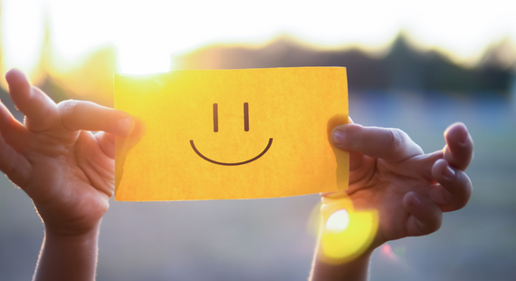 Photo of a person holding up a Post-It note with a smiley face drawn on it to camera