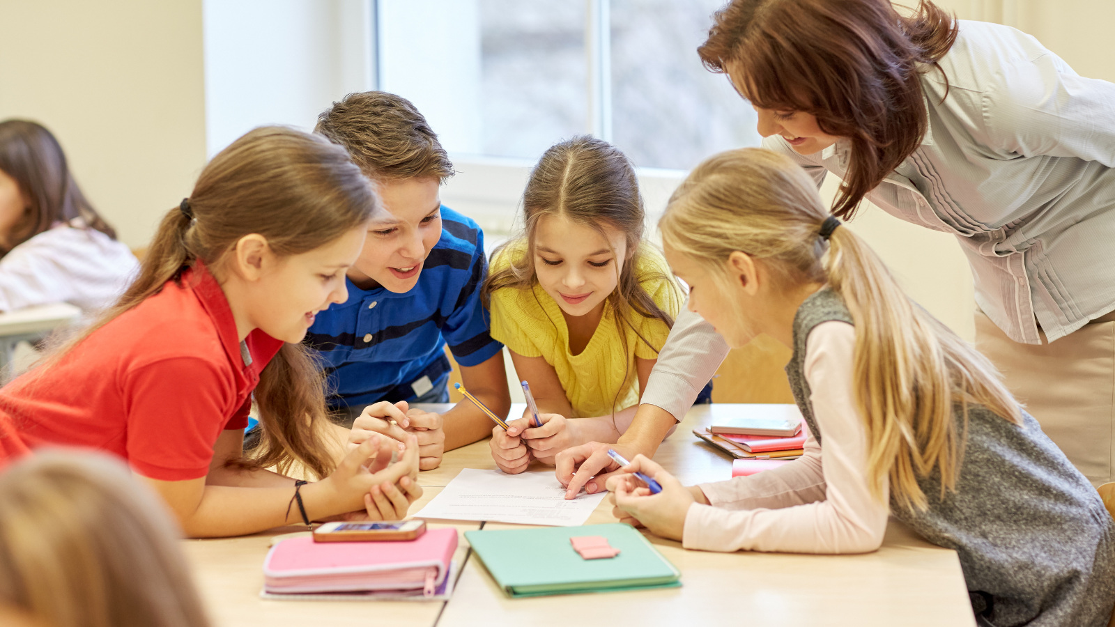 This is an image of students and teachers in a classroom gathered around workbooks and pages on a table.