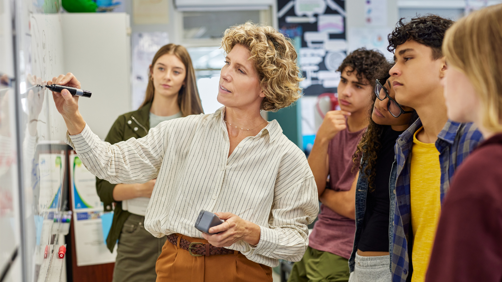 This is an image of secondary school pupils in a classroom with their teacher. The students and teacher are gathered around a white board working together on a project.