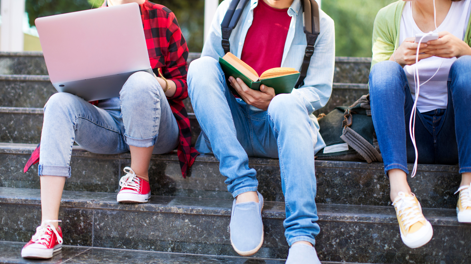 This is an image of three young people. They are sitting outside a building on steps. One of them is on a laptop, one is reading from a book and the third has a phone and headphones in their hands.